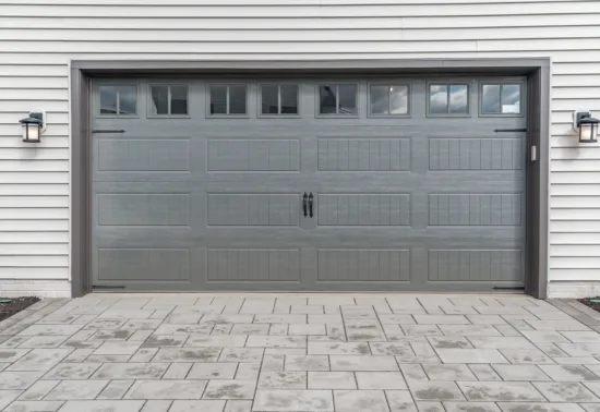 Why Is My Garage Door Not Opening Fully? AdobeStock 393448589 Gray paneled garage door with small rectangular windows at the top, flanked by two wall-mounted lantern lights, set against white siding and a patterned stone driveway—perfect for your next Illinois garage door installation.