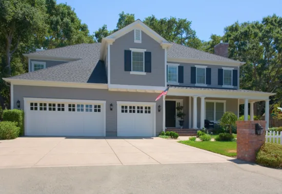 Two-story suburban house with gray siding, black shutters, three-car garage—perfect for Illinois garage door installation—front porch, an American flag, surrounded by trees and a white picket fence.