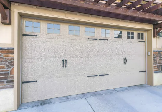 Closed beige garage door with decorative black hardware and rectangular windows along the top, set in a house exterior with stone and wood trim—perfect inspiration for your Wisconsin garage door installation or repair needs.