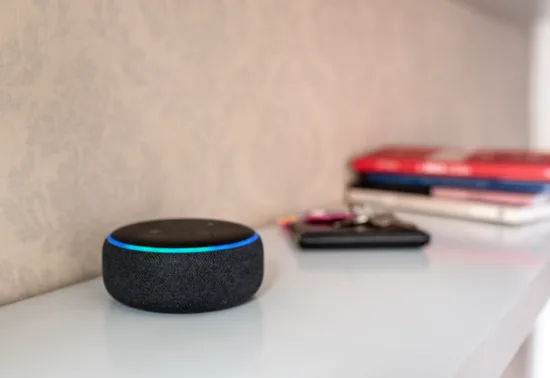 Unlock Smart Control: Connect MyQ to Alexa AdobeStock 510684015 A smart speaker with a glowing blue ring sits on a white surface next to a stack of books and other small items, perfectly blending technology with everyday essentials—much like reliable Wisconsin garage door repair keeps your home running smoothly.