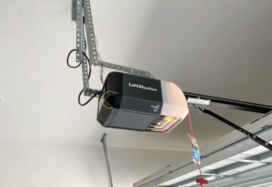Unlock Smart Control: Connect MyQ to Alexa AdobeStock 543752157 Editorial Use Only A LiftMaster garage door opener is mounted to the ceiling with metal brackets, a red emergency release cord hanging down. The partially open door showcases expert Illinois garage door installation.