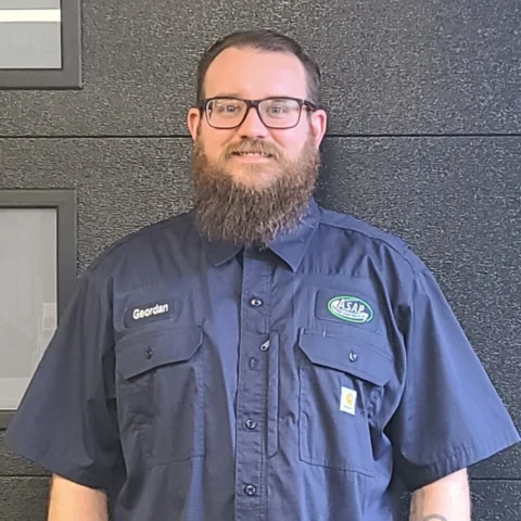 A man with glasses and a beard, wearing a navy work shirt labeled "Geordan" and an "ASAP" logo, stands in front of a black textured wall—ready for your Illinois garage door installation needs.
