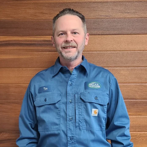 A man with short hair and a trimmed beard wears a blue work shirt with name tag "Tim" and an "ASAP" logo, standing in front of a wooden wall, representing Wisconsin garage door service professionals.