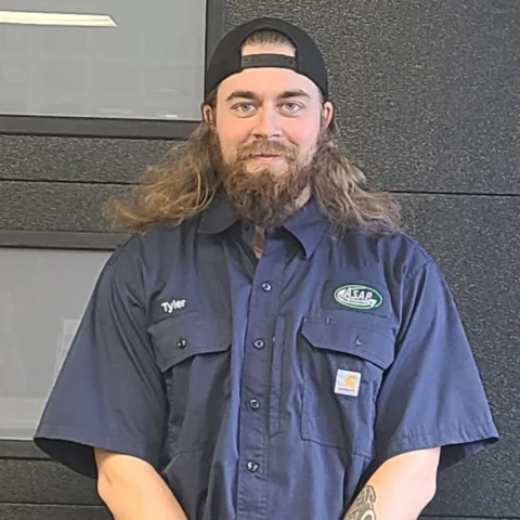 A man with long hair and a beard, wearing a backward cap, a navy work shirt with a "Tyler" name tag and an "ASAP" patch, stands in front of a dark wall—ready for Wisconsin garage door installation or service.