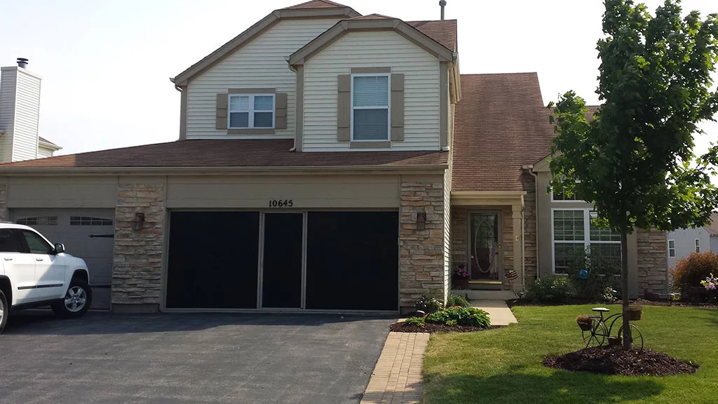 Garage Screen Doors after door image Two-story suburban house with stone and siding exterior, attached three-car garage featuring Wisconsin garage door installation, a driveway, manicured lawn, and a small tree in the front yard.