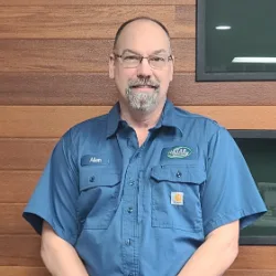 A man with glasses, a short beard, and a blue button-up shirt stands in front of a wood-paneled wall and a dark display screen, reflecting the professionalism of Wisconsin garage door service.