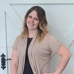 A woman with wavy, shoulder-length hair stands smiling in front of a white paneled door, wearing a beige cardigan over a dark top—perfectly showcasing the quality behind Illinois garage door service.