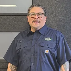 A man with glasses and tattoos wearing a navy blue button-up shirt stands smiling in front of a dark wall, representing professional Illinois garage door installation.