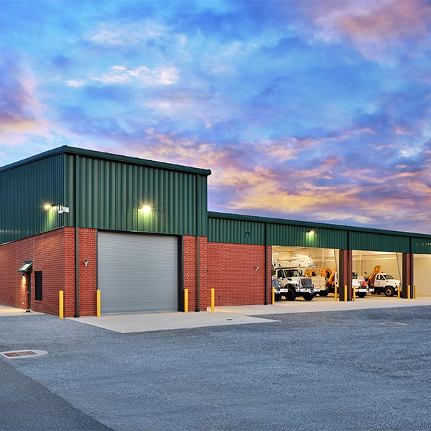 Services commercial service img A brick and metal industrial building with several open garage bays, work trucks inside, and a cloudy sunset sky in the background—ideal for businesses seeking reliable Illinois garage door repair or installation services.