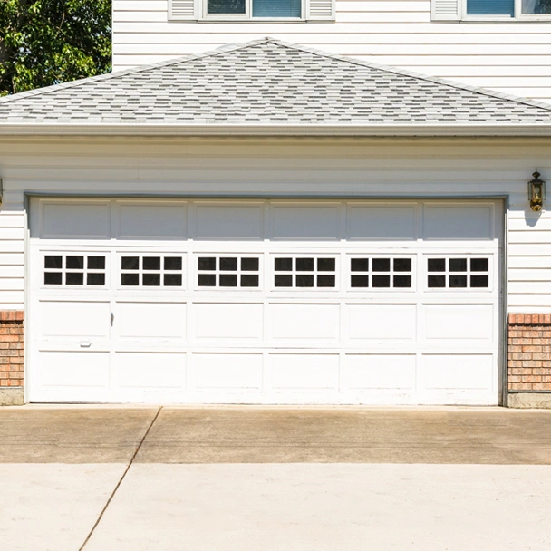 Services custom garage door design img A closed white double garage door with small square windows, attached to a house with white siding and partial brickwork, showcases quality Illinois garage door installation on a sunny day.