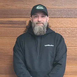 A man with a long beard, wearing a black hoodie and cap, stands in front of a wooden wall—perfectly capturing the reliable look of an Illinois garage door installation specialist.
