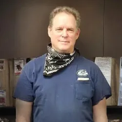 A man wearing a blue shirt and a patterned scarf stands indoors in front of a dark background with paper bags and papers posted behind him, reminiscent of an Illinois garage door installation workspace.