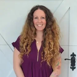A woman with long curly hair wearing a purple top stands in front of a white barn-style door, smiling at the camera—showcasing the quality of Illinois garage door installation.