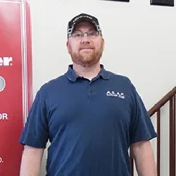 A man with a beard and glasses wearing a navy blue polo shirt and black cap stands indoors near a red sign and a staircase, representing Illinois garage door installation.