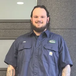 Man with dark hair and beard wearing a dark blue work shirt stands in front of a textured wall and glass panel, representing a trusted Wisconsin garage door service professional.