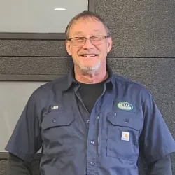 A man wearing glasses and a dark blue work shirt with a name tag stands in front of a gray textured wall, smiling at the camera—ready to provide expert Illinois garage door installation.