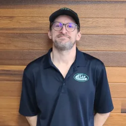 Man wearing glasses, a black cap, and a black ASAP polo shirt stands in front of a wooden wall, representing Wisconsin garage door installation, facing the camera with hands behind his back.