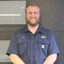 A man with a beard wearing a navy blue work shirt that says "Tyler" stands smiling in front of a dark wall and glass panels, ready to tackle any Illinois garage door repair with expertise.