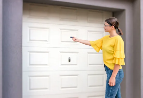 A woman wearing a yellow top and jeans stands in front of a closed garage door, pointing a remote control at it, possibly testing the work done by a garage door company Elgin residents trust.
