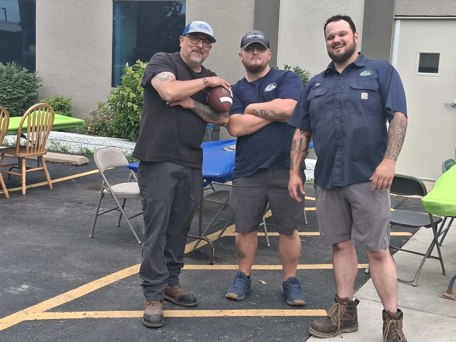 Recognizing Our Incredible Technicians tech day Three men with tattoos stand together outdoors in a parking lot near tables and chairs; one is holding a football.