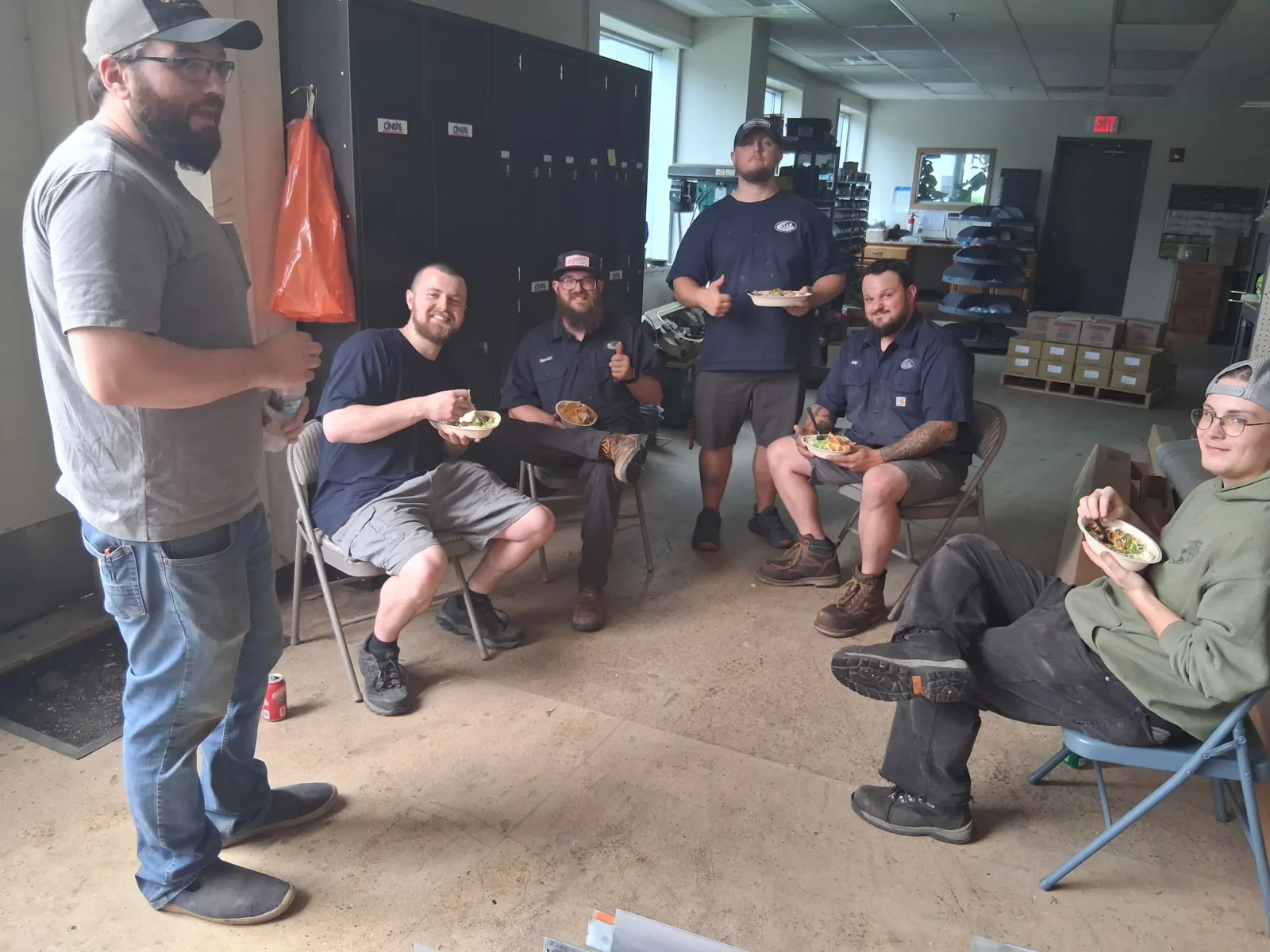 Recognizing Our Incredible Technicians tech day 2 Six men sit and stand in a break room, eating food and smiling. The room contains lockers, shelves, and work supplies. Casual, relaxed atmosphere.