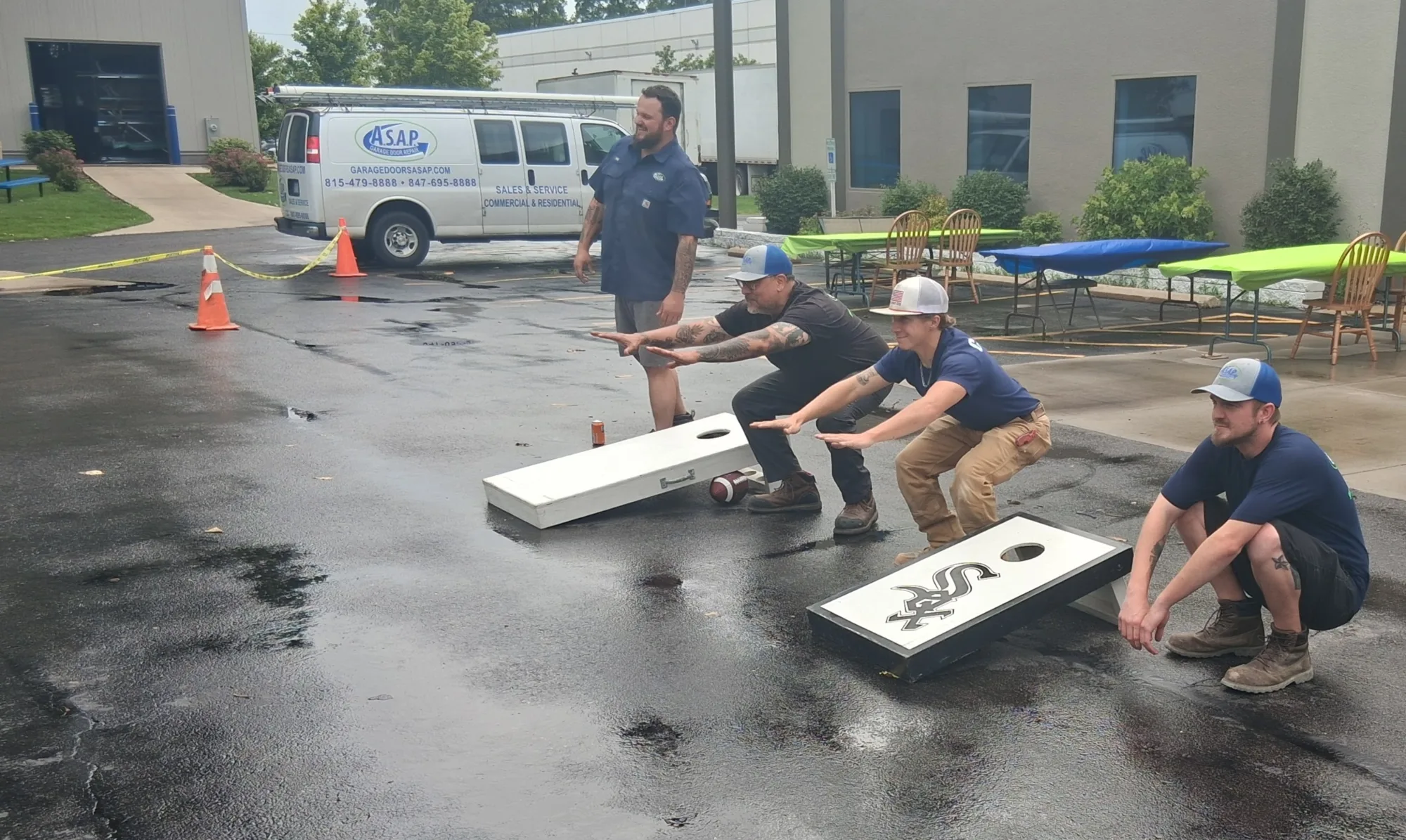 Recognizing Our Incredible Technicians tech day 3 Four people in work uniforms squat in front of cornhole boards on wet pavement near a building, with a white van and outdoor tables in the background.
