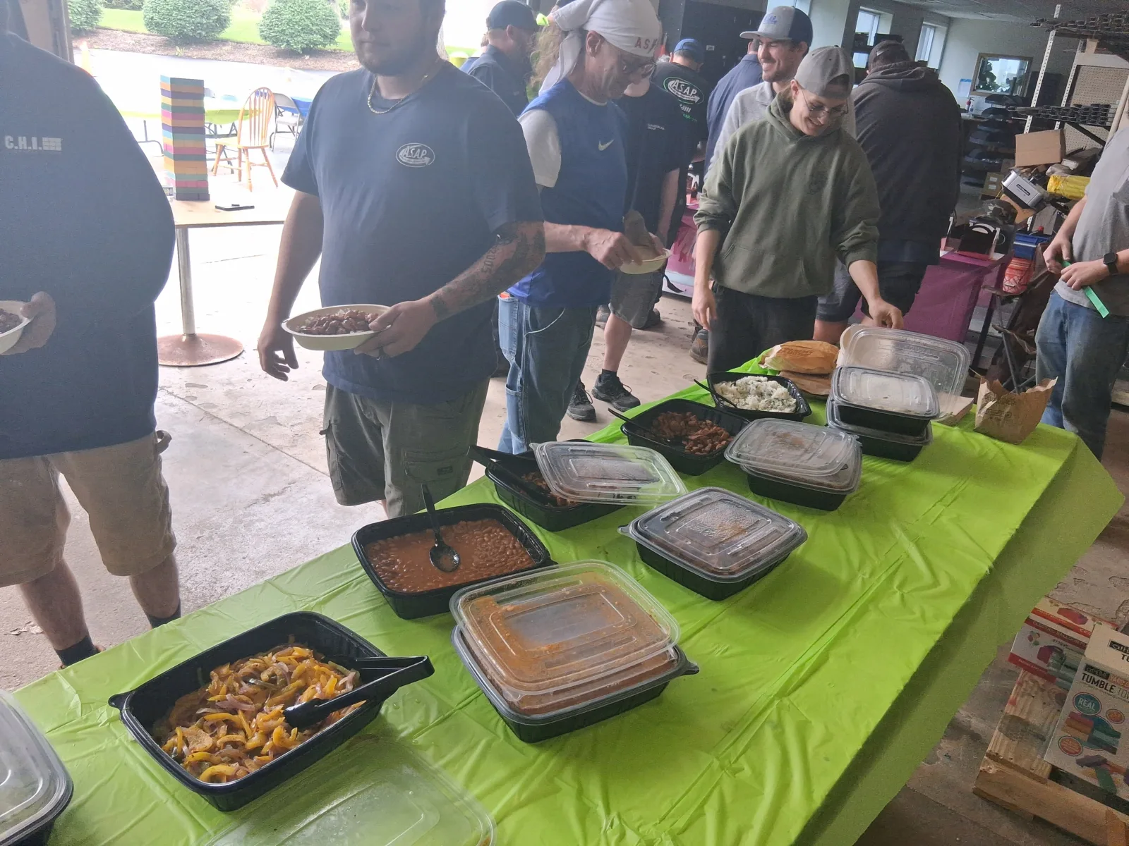 Recognizing Our Incredible Technicians tech day 6 Several people stand in line at a buffet table with various covered and uncovered food trays on a green tablecloth in a casual indoor setting.